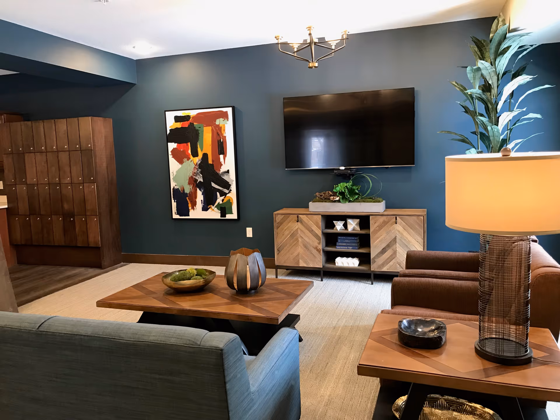Modern communal living room with a teal accent wall, wall-mounted TV, wooden media cabinet, seating, coffee tables, artwork and a floor lamp.