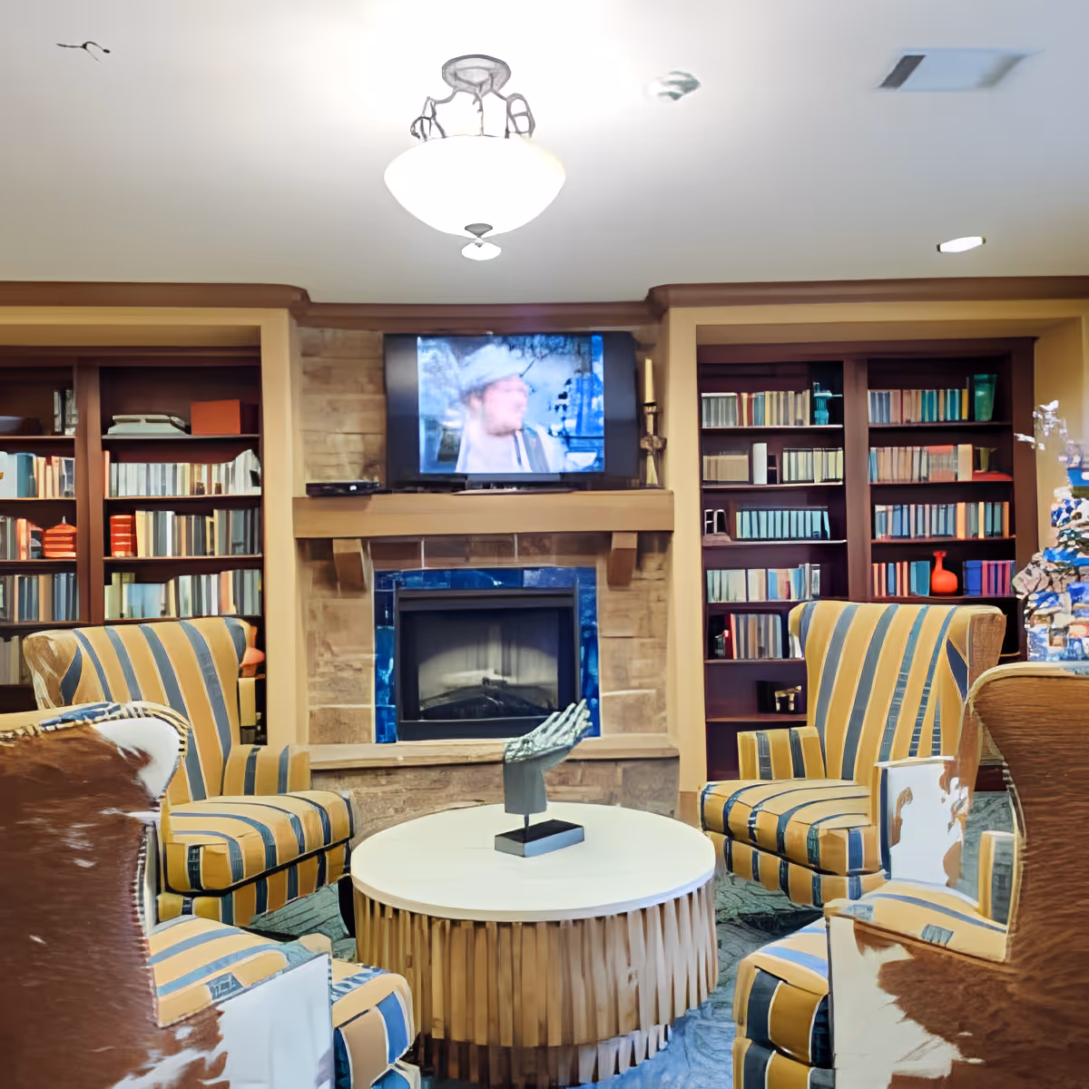 A cozy living room area with four striped armchairs arranged around a round wooden coffee table. Behind the seating area is a stone fireplace with a flat-screen TV mounted above it. On either side of the fireplace are built-in bookshelves filled with books and decorative items. The room is warmly lit with ceiling lights.