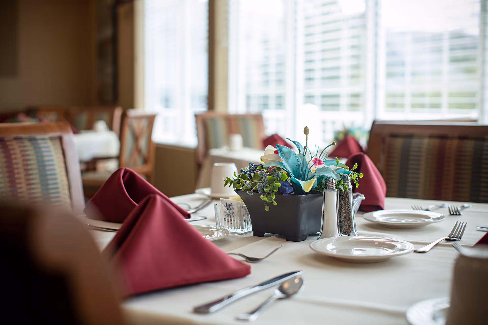 A dining table set with white plates, silverware, and burgundy folded napkins. A black rectangular vase with artificial flowers and salt and pepper shakers are placed in the center of the table. The background shows more tables and chairs near large windows with blinds.