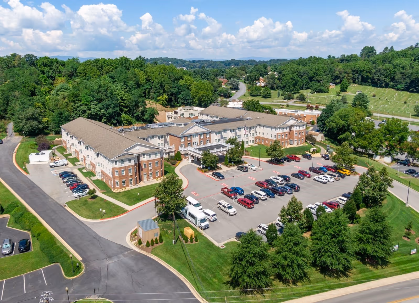Aerial view of The Legacy at North Augusta senior living facility showing a large multi-wing building with a parking lot filled with cars in front, surrounded by green trees and grassy areas under a partly cloudy sky.
