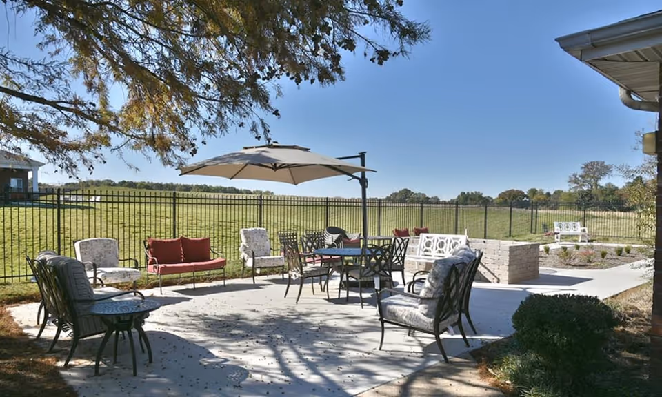 Outdoor patio seating area with chairs, tables, a large umbrella and a firepit overlooking a fenced grassy field.