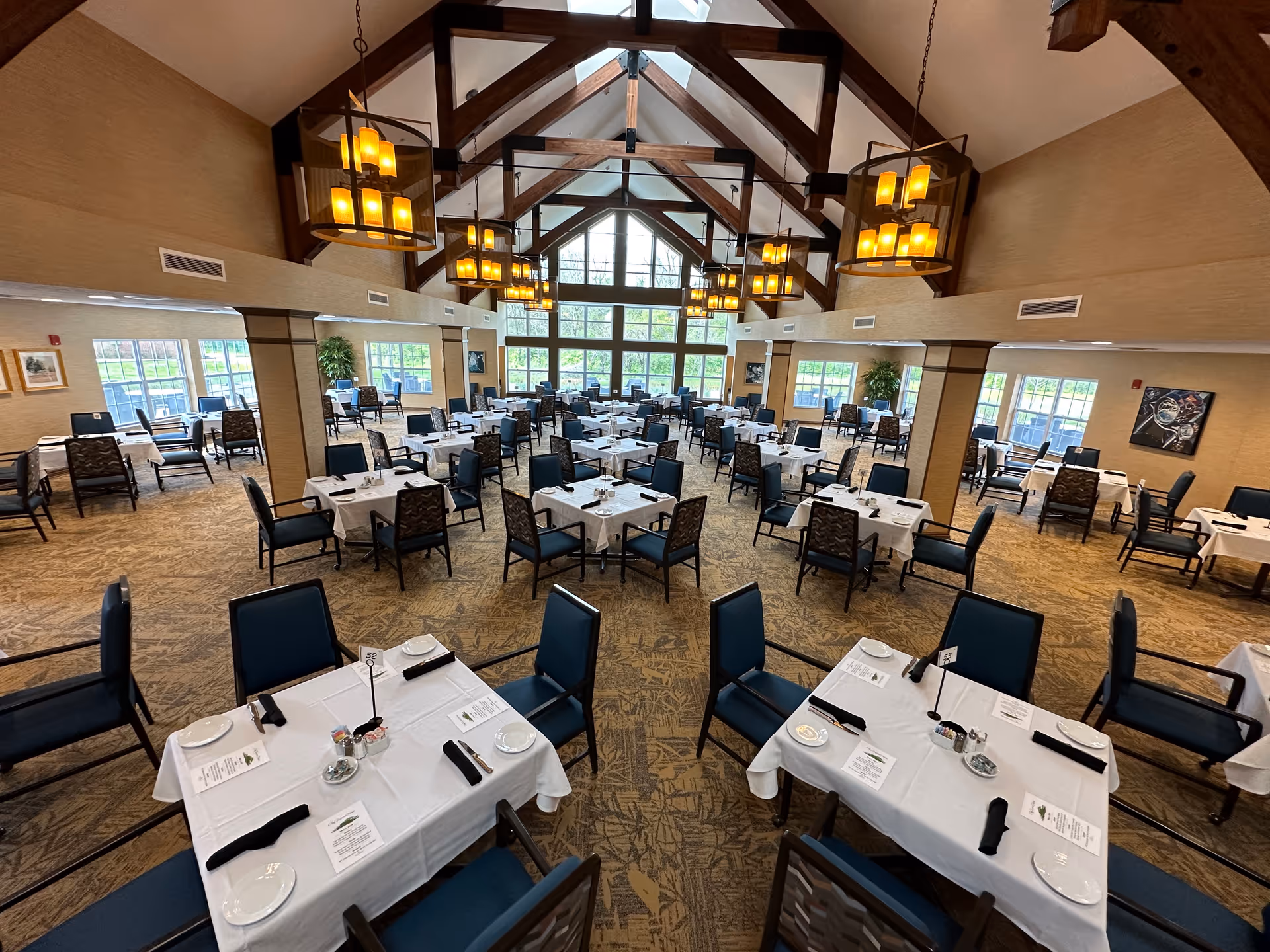 Spacious dining room with multiple tables covered in white tablecloths, each set with plates, napkins, menus, and condiments. The room features high vaulted ceilings with exposed wooden beams and large hanging light fixtures. Large windows at the far end allow natural light to fill the space, and there are several chairs arranged around each table.