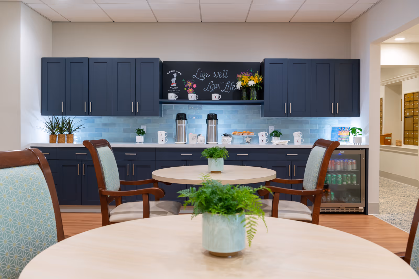 A cozy kitchen area with dark blue cabinets and a light blue tiled backsplash. There are two round tables with wooden chairs that have light blue patterned cushions. On the tables are small green potted plants. The countertop holds coffee dispensers, mugs, a plate of pastries, and decorative plants. Above the counter is a chalkboard with the phrase 'Live well Love Life' written on it, along with a small illustration of a dog and the words 'Good Dog Cafe'.