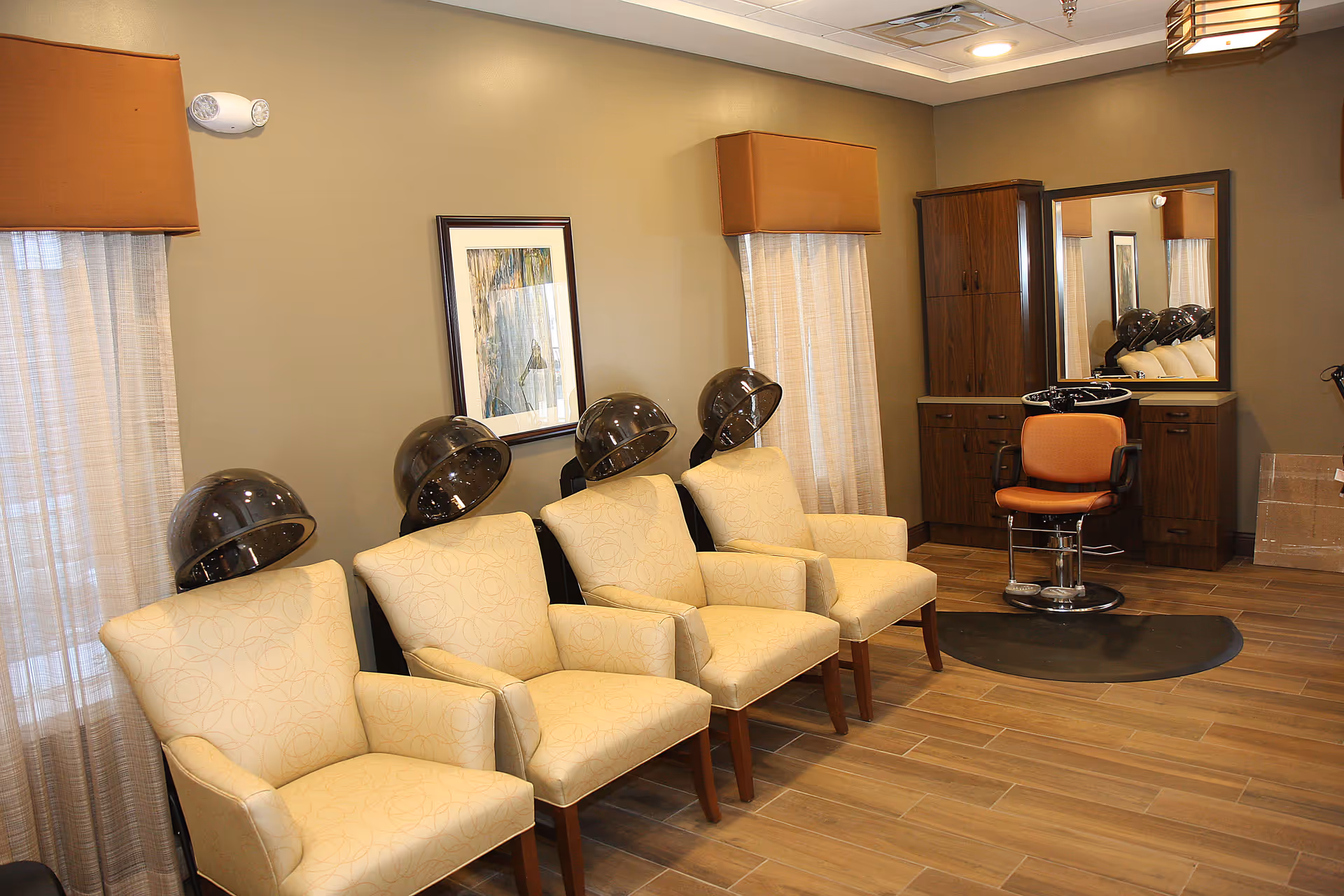 Interior view of a salon area with four beige armchairs each positioned under a hair dryer hood. There is a framed artwork on the wall, two windows with curtains and valances, and a wooden cabinet with a mirror and an orange salon chair in front of a hair washing station.