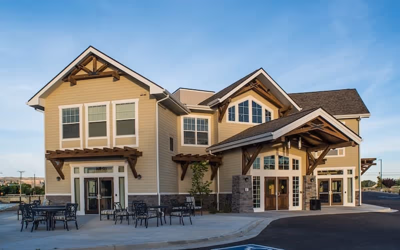 Exterior view of a two-story senior living facility building with beige siding, large windows, and wooden architectural accents. There are outdoor tables and chairs on a concrete patio in front of the building under a clear blue sky.