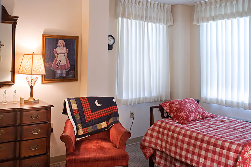 A cozy bedroom with a single bed covered in red and white checkered bedding and a matching pillow. Next to the bed is a red armchair draped with a quilt featuring moon and star patterns. A wooden dresser with a lamp and decorative items sits against the wall, and a framed picture of a doll hangs above it. Two windows with white curtains allow natural light into the room.