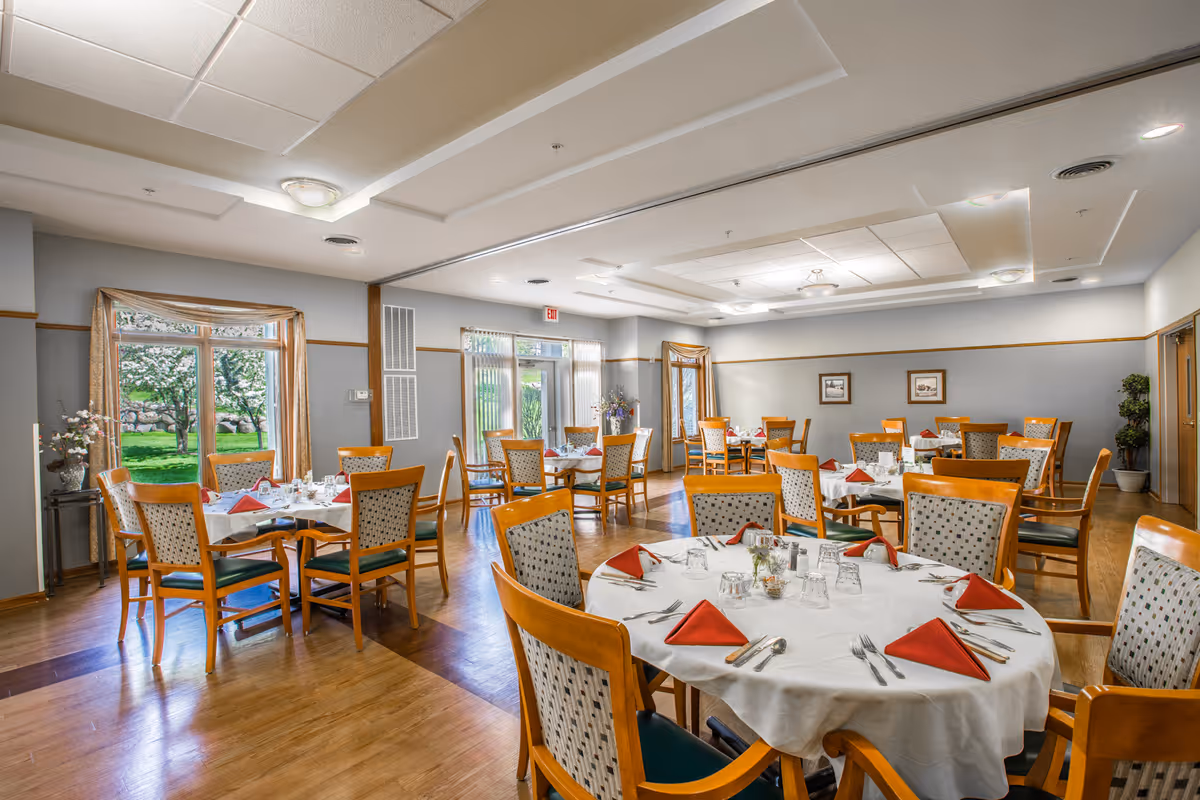 A bright dining room with multiple round tables covered in white tablecloths, each set with red folded napkins, glasses, and silverware. Wooden chairs with patterned cushions surround the tables. Large windows and glass doors allow natural light to fill the room, and there are plants and framed pictures on the walls.