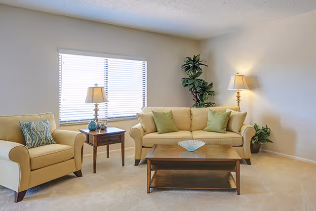 A bright living room with beige walls and carpet, featuring a beige sofa with green throw pillows, a matching armchair with a patterned pillow, a wooden coffee table, two table lamps on wooden side tables, and two green plants in the corners.