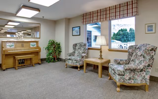 A cozy sitting area in a senior living facility featuring two floral upholstered armchairs with wooden legs, a wooden side table with a lamp, a wooden piano with a matching bench, a potted plant, and two windows with plaid valances showing an outdoor view.