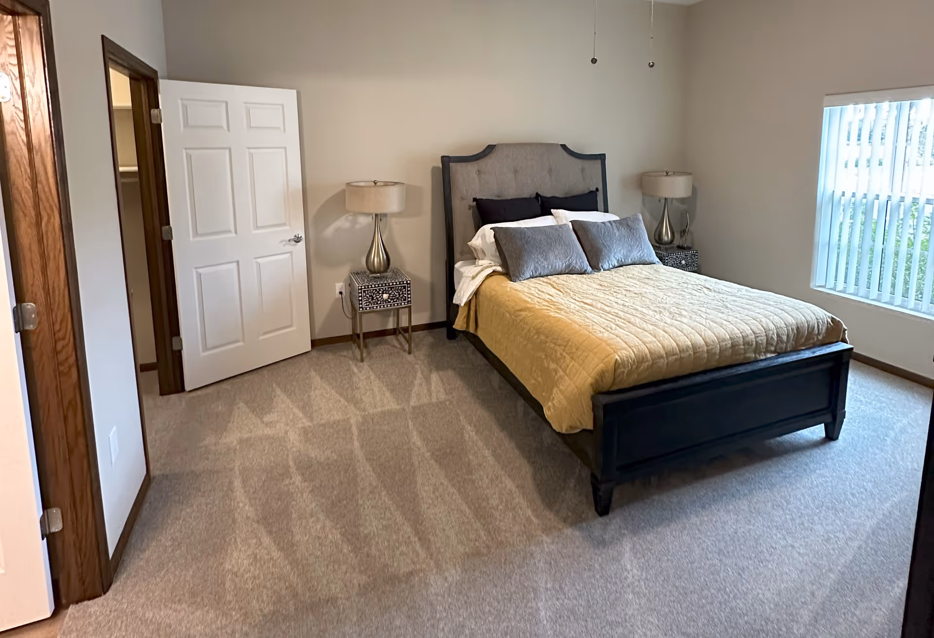 Bright, simply furnished bedroom with a bed covered by a yellow quilt, two bedside tables with lamps, an open closet door and a window with vertical blinds.