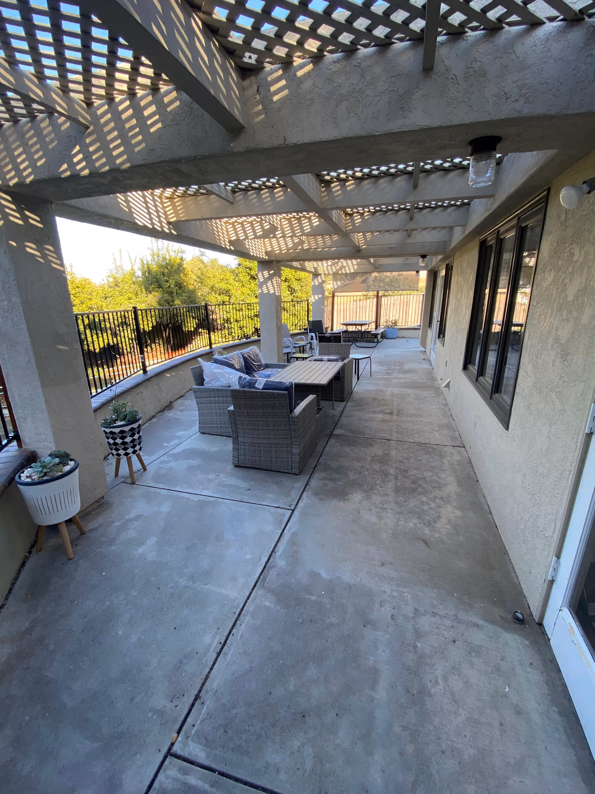 Outdoor covered patio area with concrete floor and lattice roof casting shadows. The patio is furnished with wicker seating including chairs and a sofa with cushions, coffee tables, and potted plants on stands. A black metal fence borders the patio with greenery visible beyond.