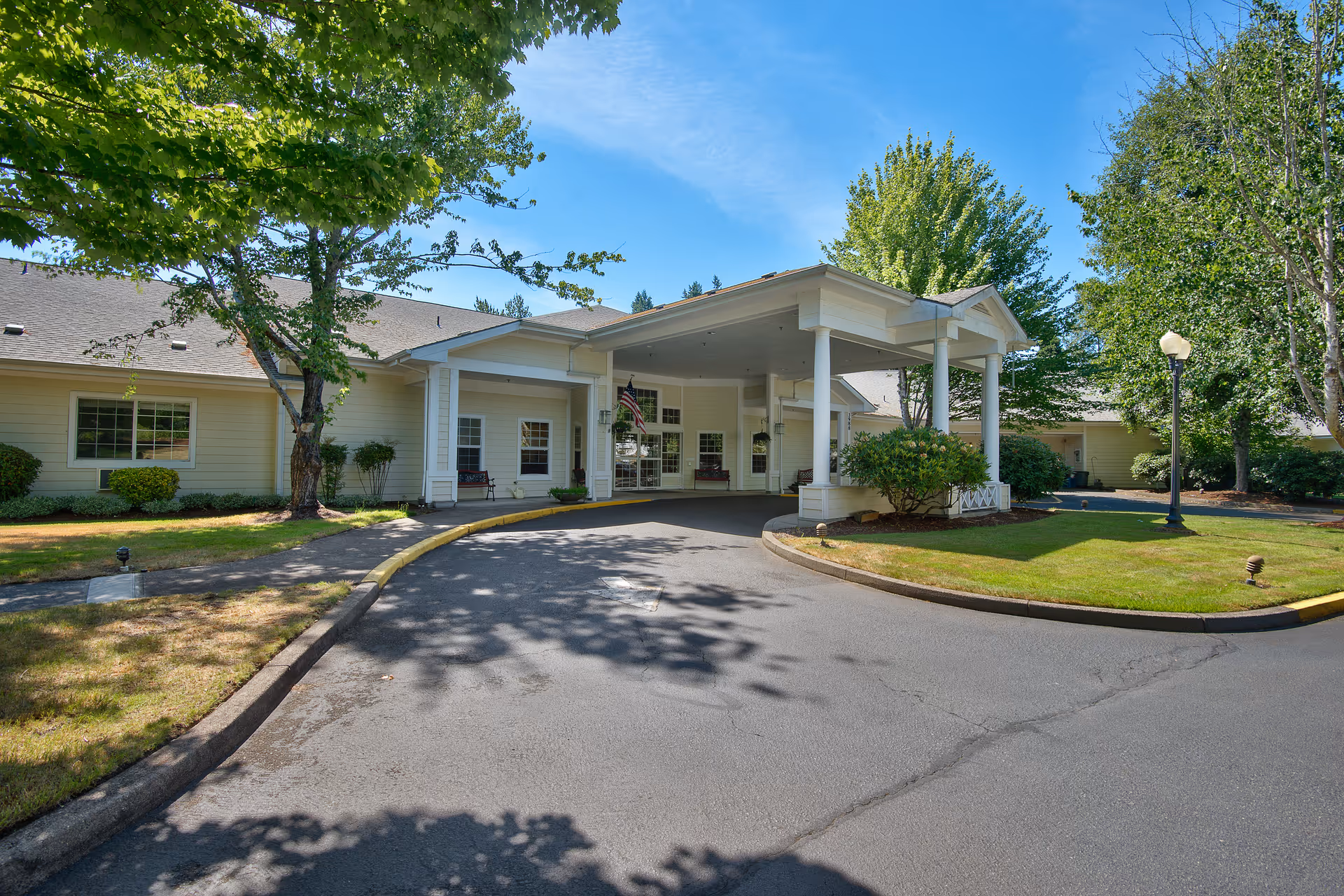 Entrance of Meadow Creek Village Assisted Living facility showing a covered drop-off area with white pillars, surrounded by green trees and well-maintained landscaping under a clear blue sky.