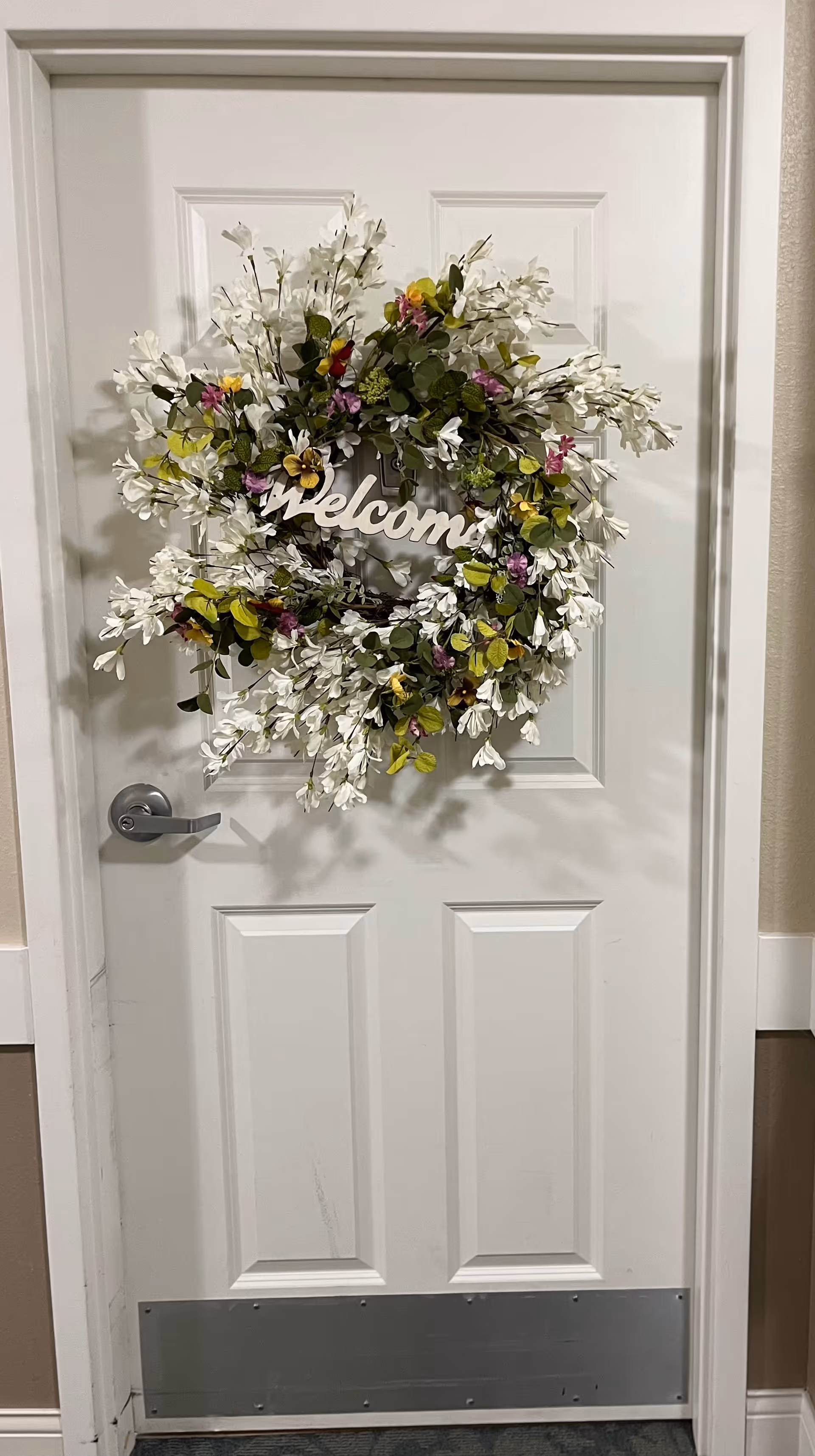 A white door with a silver handle and a metal kick plate at the bottom. Hanging on the door is a decorative wreath made of white flowers and green leaves with some small purple and yellow flowers, and a wooden sign in the center that says 'welcome'.