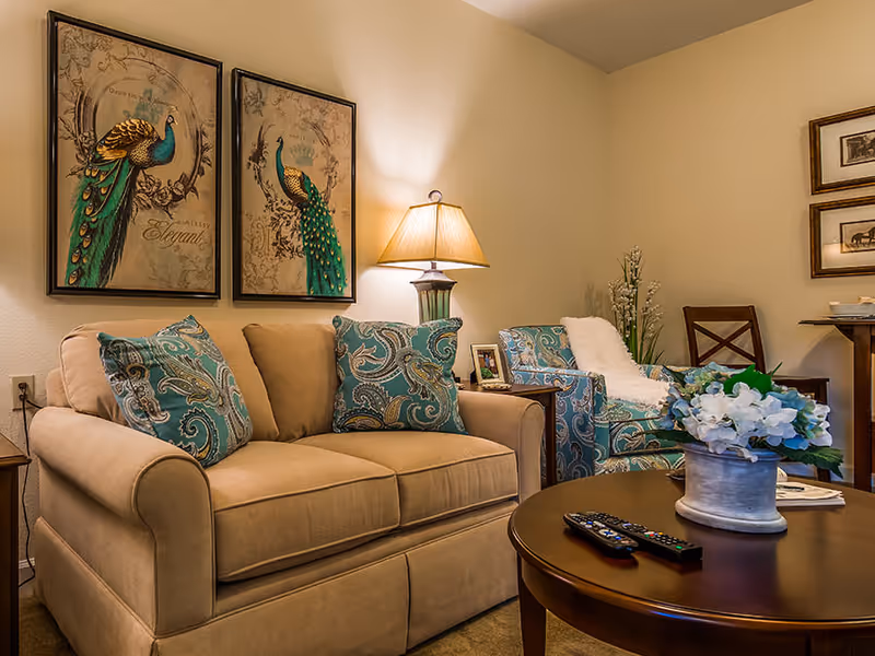 Cozy living room with a beige sofa, patterned armchair, round coffee table with remotes and a floral arrangement, a lamp, and peacock wall art.