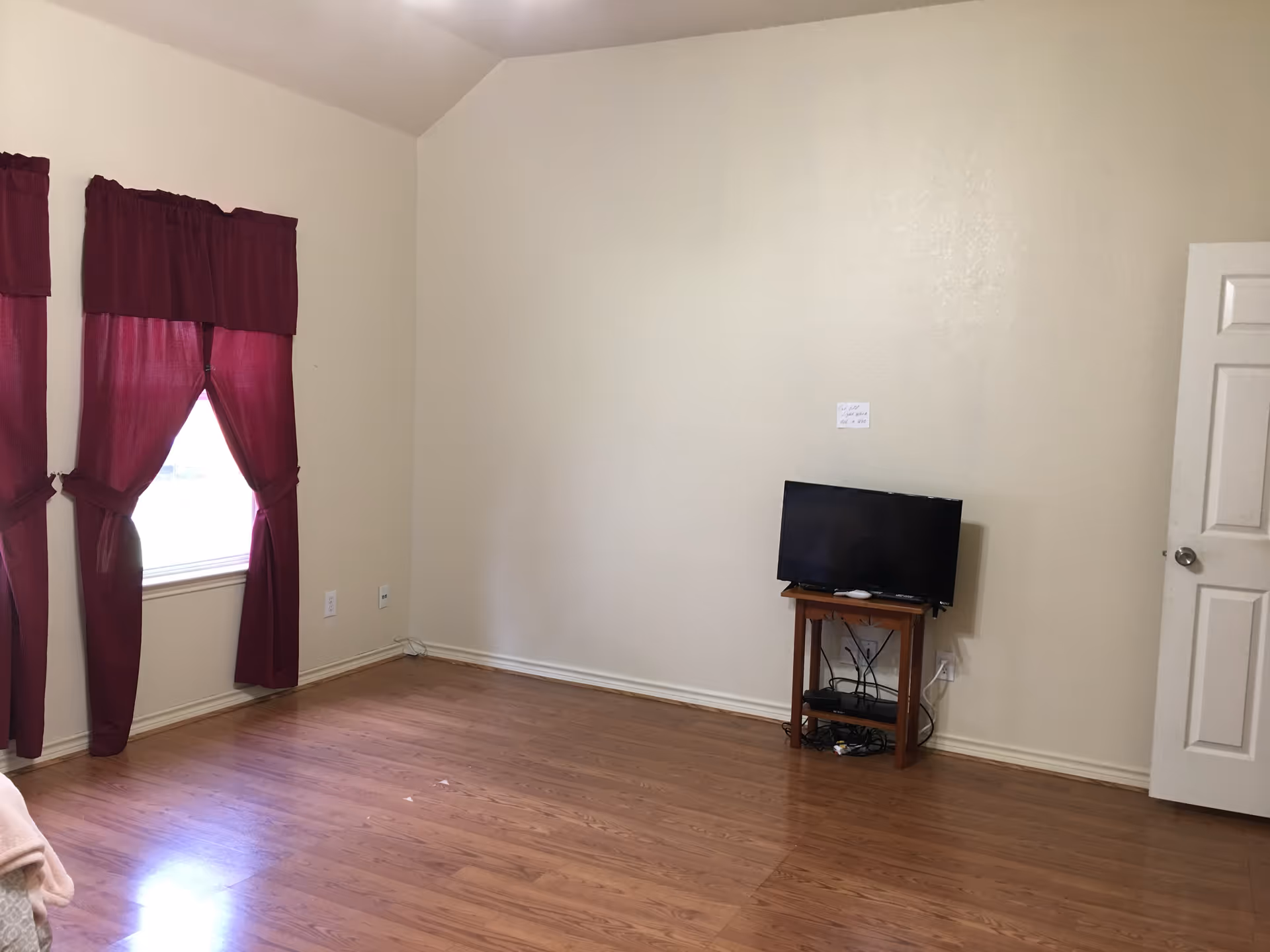 A sparsely furnished room with wooden flooring, cream-colored walls, and a window with dark red curtains. A small wooden stand holds a flat-screen TV and some electronic devices. A white door is partially open on the right side of the image.