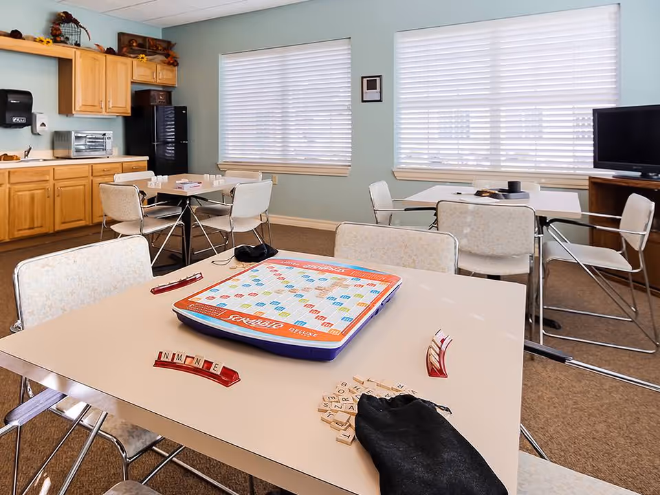 Well-lit common activity/dining room with tables and chairs and a Scrabble game set up on the nearest table.