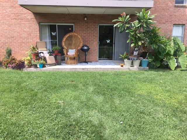 Outdoor patio area of an apartment with a brick exterior wall, sliding glass doors, various potted plants, a wicker chair with a cushion, a reclining chair, and a small charcoal grill, all overlooking a well-maintained grassy lawn.