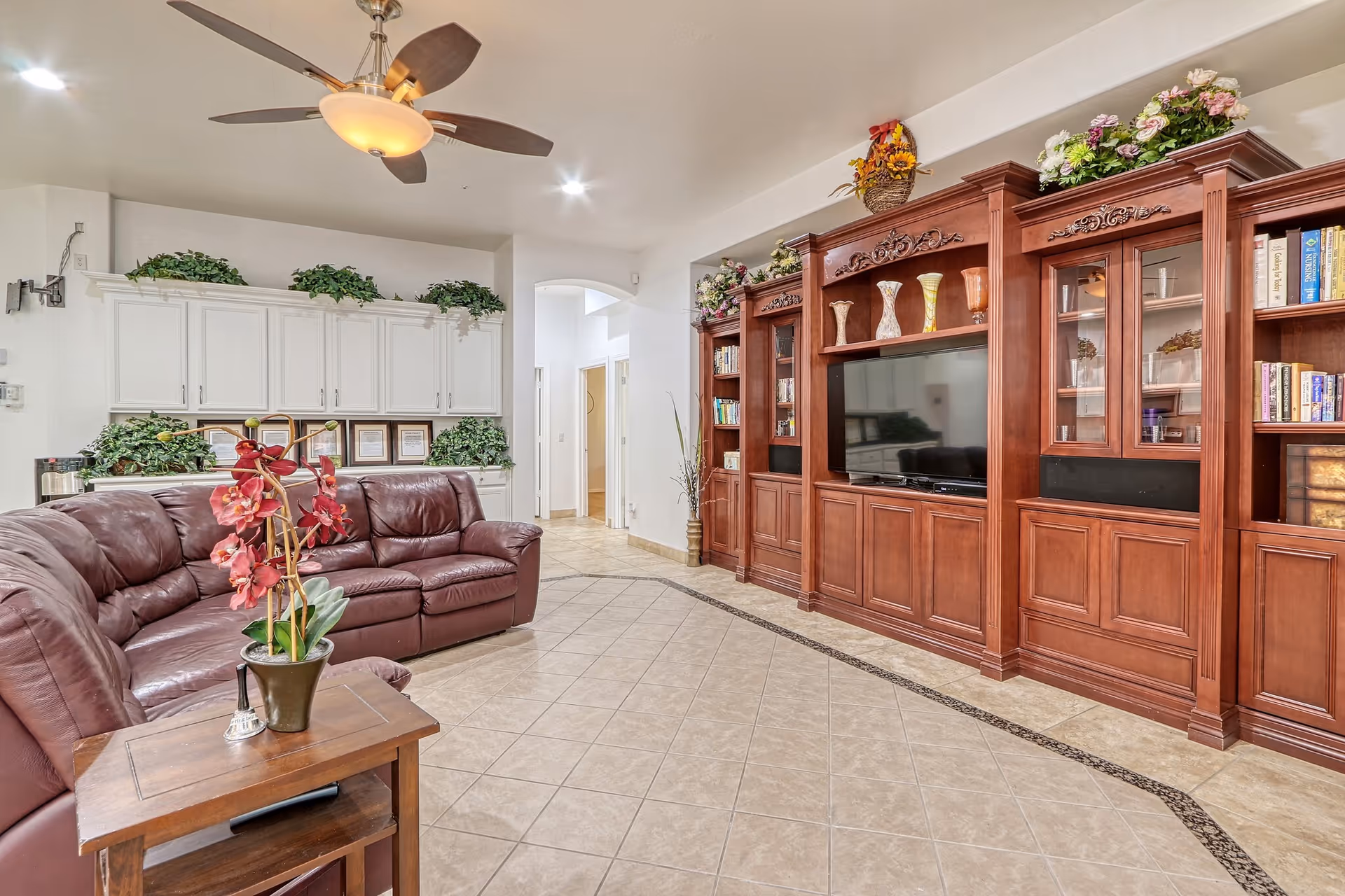 A spacious living room with a large brown leather sectional sofa, a wooden side table with a potted orchid plant, a ceiling fan with light, and a large wooden entertainment center with a flat-screen TV and decorative vases. The room has tiled floors and white walls with built-in white cabinets and green plants on top.