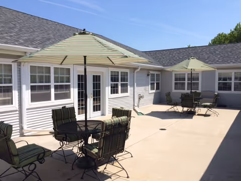 Outdoor patio area at Wisteria Haus with two round tables, each surrounded by four cushioned chairs with green striped fabric. Each table has a large green and white striped umbrella providing shade. The patio is adjacent to a light gray building with multiple windows and a glass door. The sky is clear and blue.