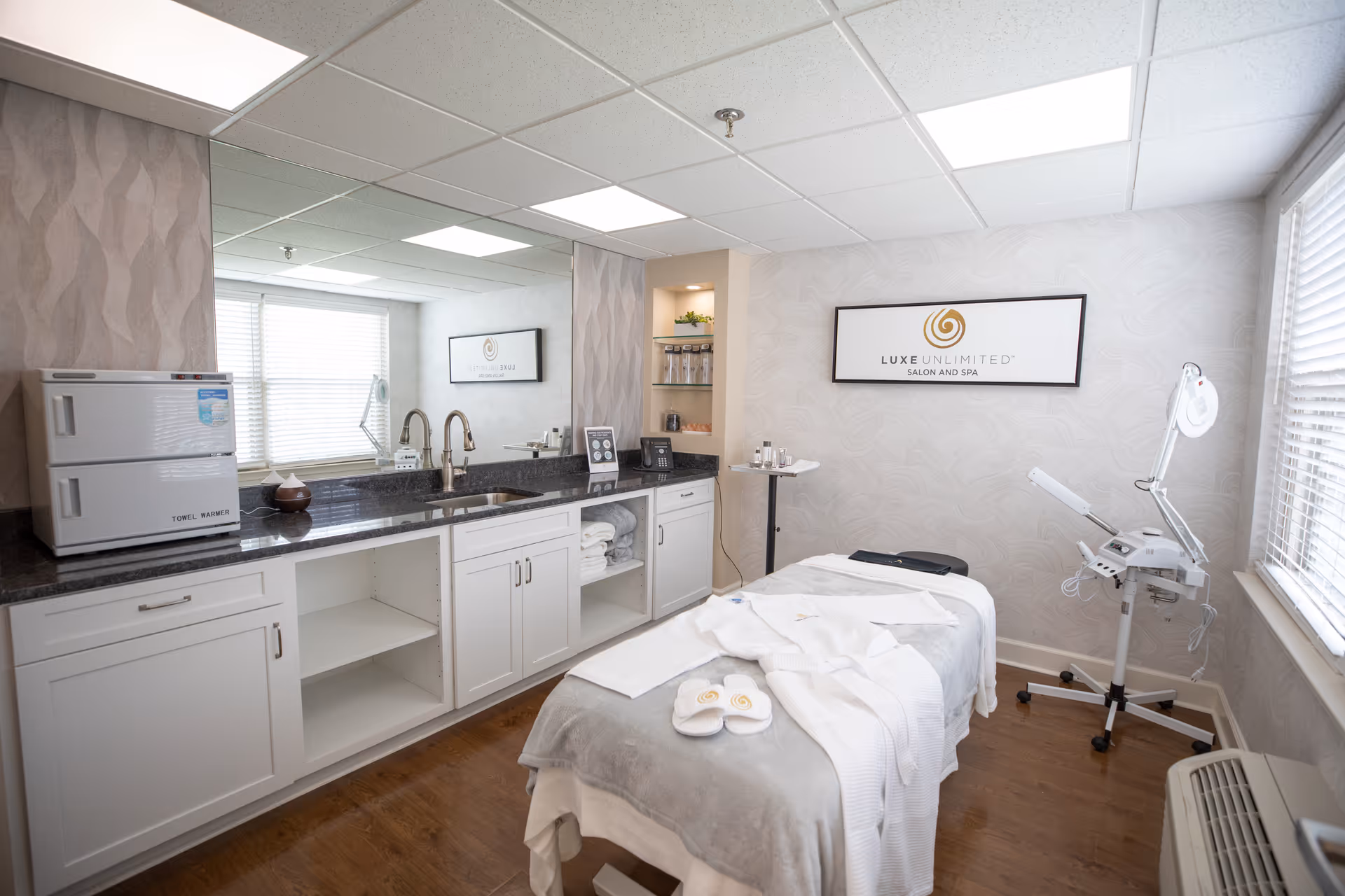 A bright and clean spa treatment room with a massage table covered in white towels and slippers. The room features a large mirror above a countertop with a sink, towel warmer, and various spa products. A sign on the wall reads 'Luxe Unlimited Salon and Spa'. The room has wood flooring and large windows with blinds letting in natural light.