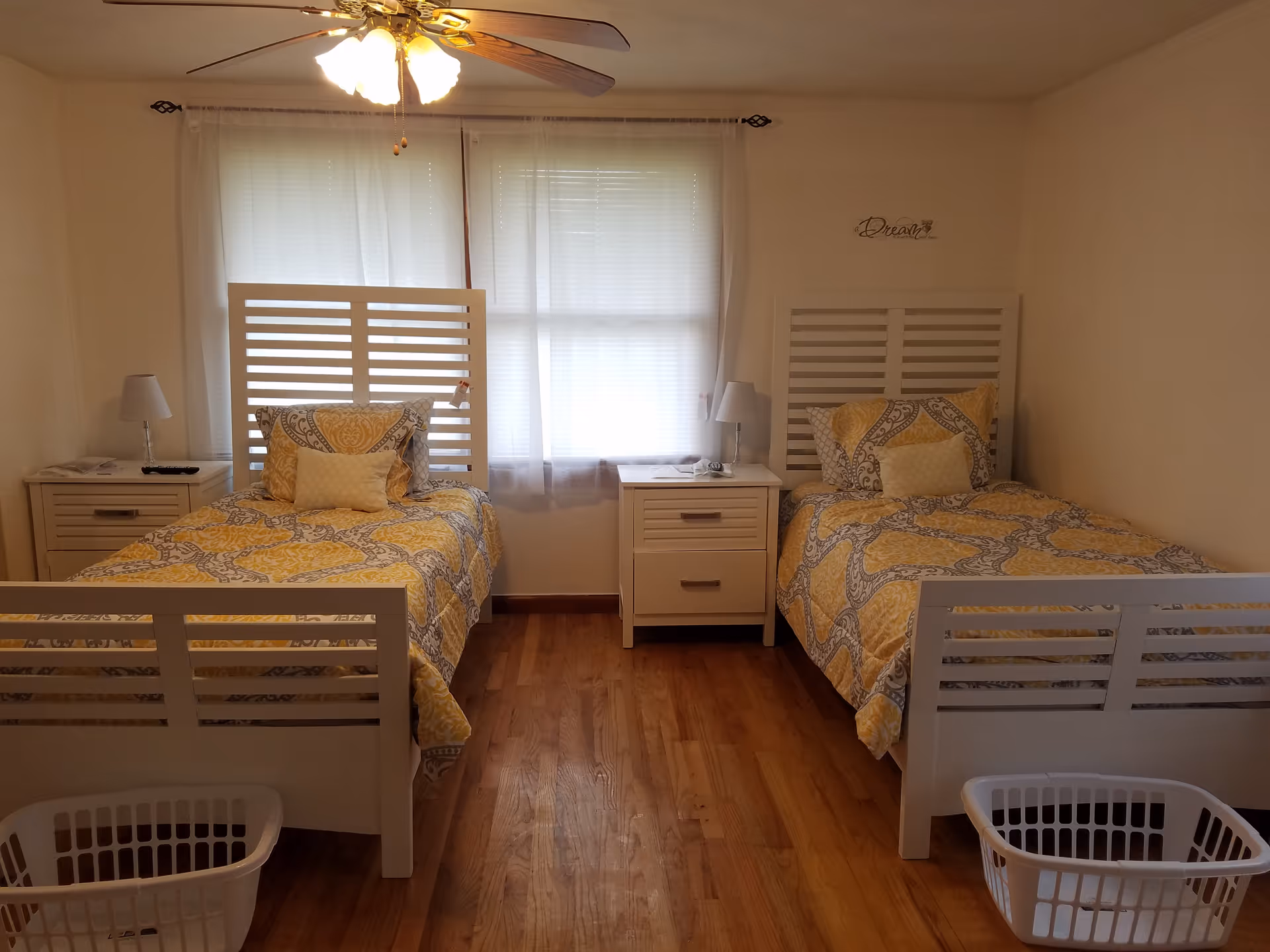 Two single beds with yellow patterned bedding in a tidy room with a central nightstand, ceiling fan, and laundry baskets at the foot of each bed.