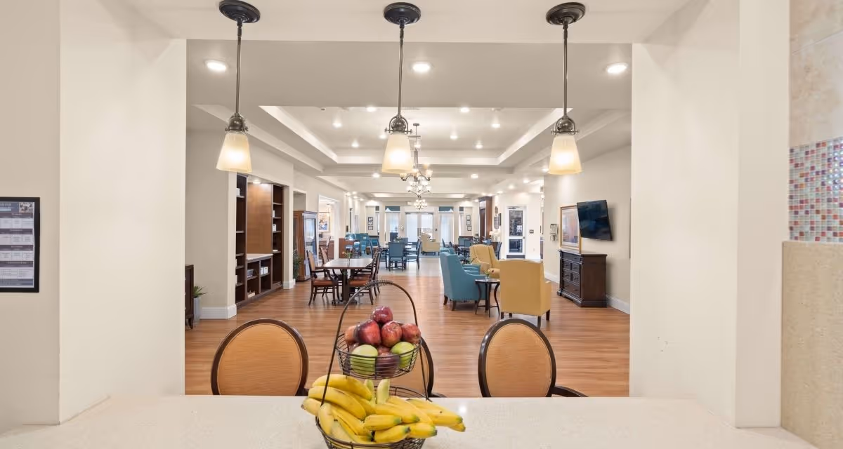 View from a kitchen counter looking into a spacious common area with wooden floors, multiple seating arrangements including tables with chairs and armchairs in blue and yellow. The ceiling has recessed lighting and hanging pendant lights. A basket of bananas and apples is placed on the counter in the foreground.