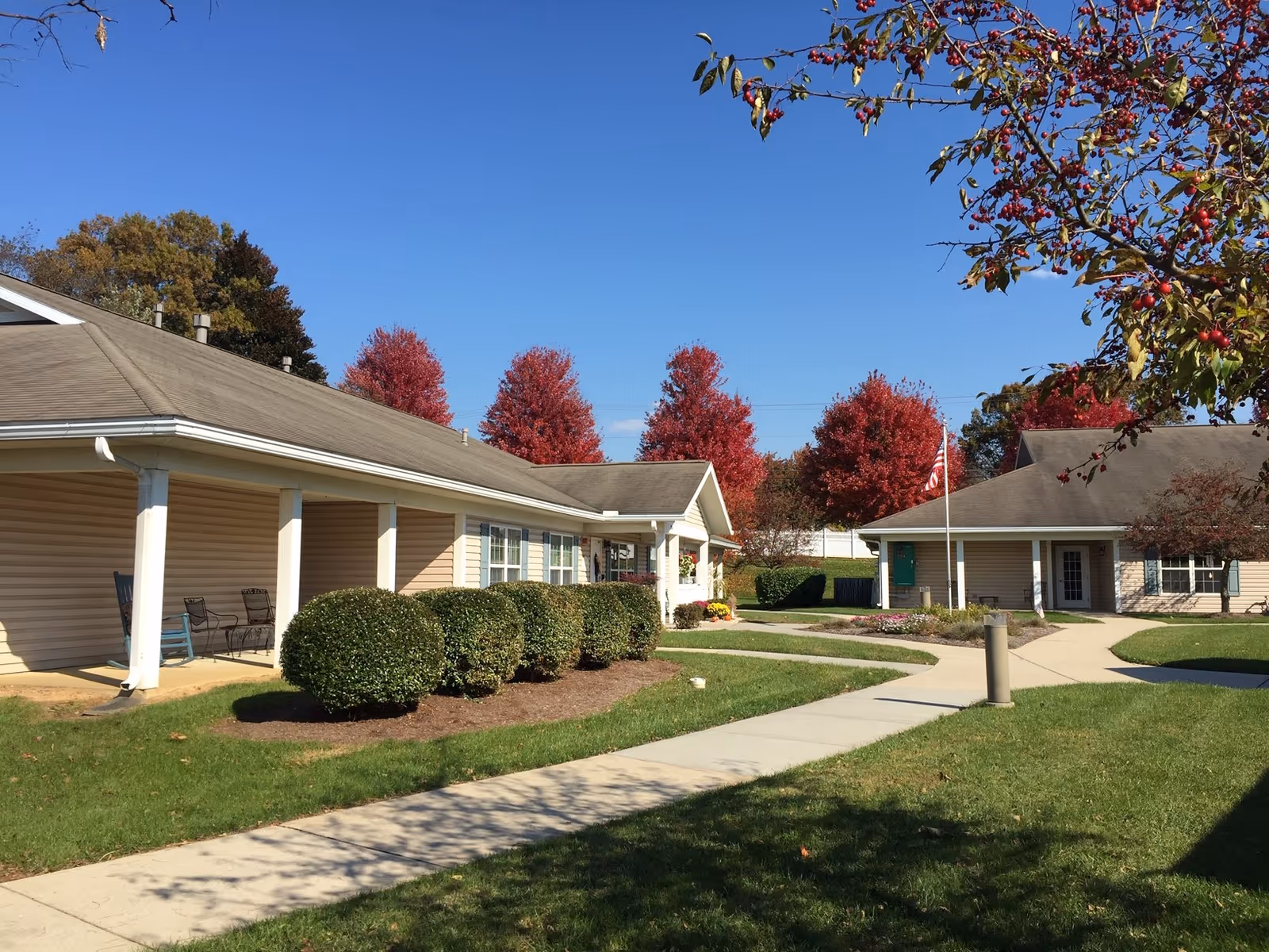 Single-story senior living community buildings flanking a paved walkway with trimmed shrubs, fall-colored trees and an American flag.