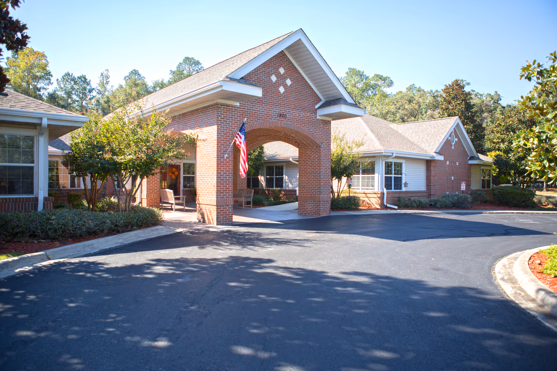 Front exterior view of a brick building with a covered entrance featuring an archway and an American flag. There are benches under the covered area and trees and shrubs surrounding the building. The sky is clear and blue.