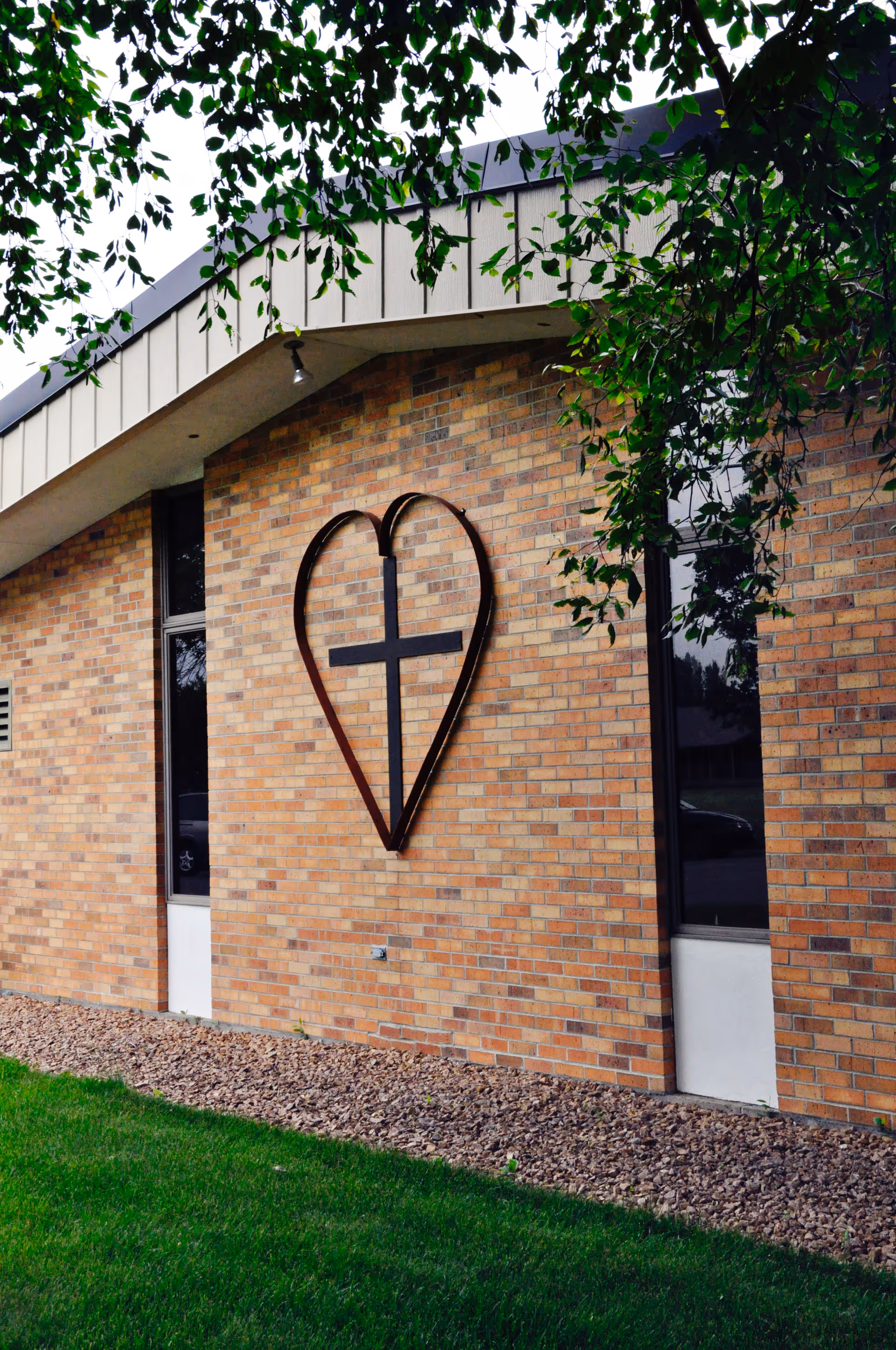 Exterior brick wall of a building with a metal heart-shaped frame enclosing a cross mounted on it, green grass and tree branches partially visible.