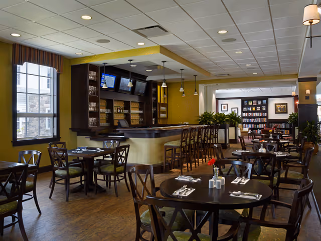 Interior view of a dining area in The Heritage of Green Hills featuring multiple round tables with chairs, each set with napkins and silverware. There is a bar counter with high stools and shelves stocked with glasses and bottles. The room has large windows, pendant lights, and a cozy atmosphere with warm yellow walls and wooden flooring. In the background, there is a lounge area with bookshelves and framed artwork.
