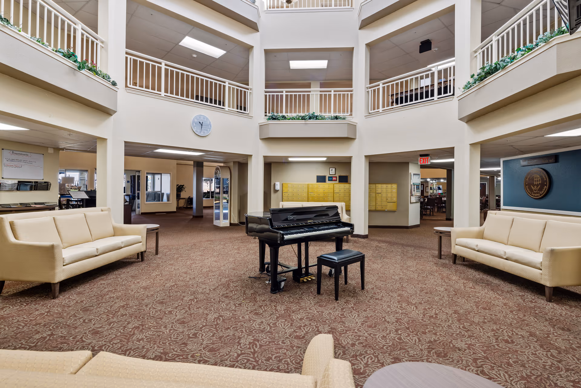 Spacious senior living community common area with a grand piano and piano bench in the center, surrounded by beige sofas and small round tables. The room has a patterned carpet, cream-colored walls, and a high ceiling with multiple balconies overlooking the space. A clock is mounted on the wall above the piano, and mailboxes are visible in the background.