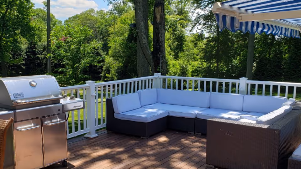 Outdoor wooden deck with a stainless steel grill on the left and a large L-shaped wicker patio sofa with light blue cushions on the right. The deck is surrounded by a white railing and overlooks a green, tree-filled yard under a partly cloudy sky. A blue and white striped awning extends over part of the seating area.