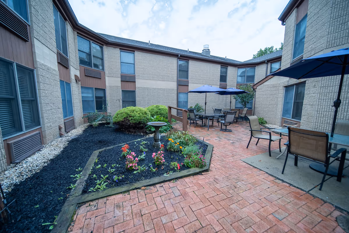 Outdoor courtyard area of a senior living facility with brick paving, garden beds with flowers and shrubs, several tables with chairs, and blue umbrellas providing shade. The courtyard is surrounded by a two-story building with multiple windows.