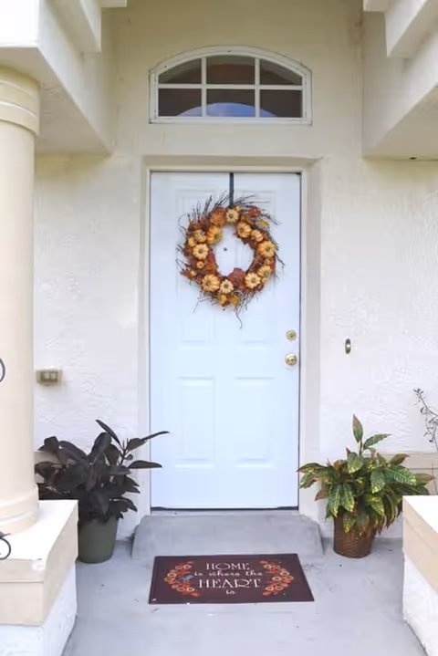 Front entrance of a house with a white door decorated with a fall-themed wreath made of small pumpkins and flowers. There are two potted plants on either side of the door, one with dark leaves and the other with green and yellow leaves. A doormat in front of the door reads 'Home is where the heart is.' The exterior walls are light-colored with a small arched window above the door.