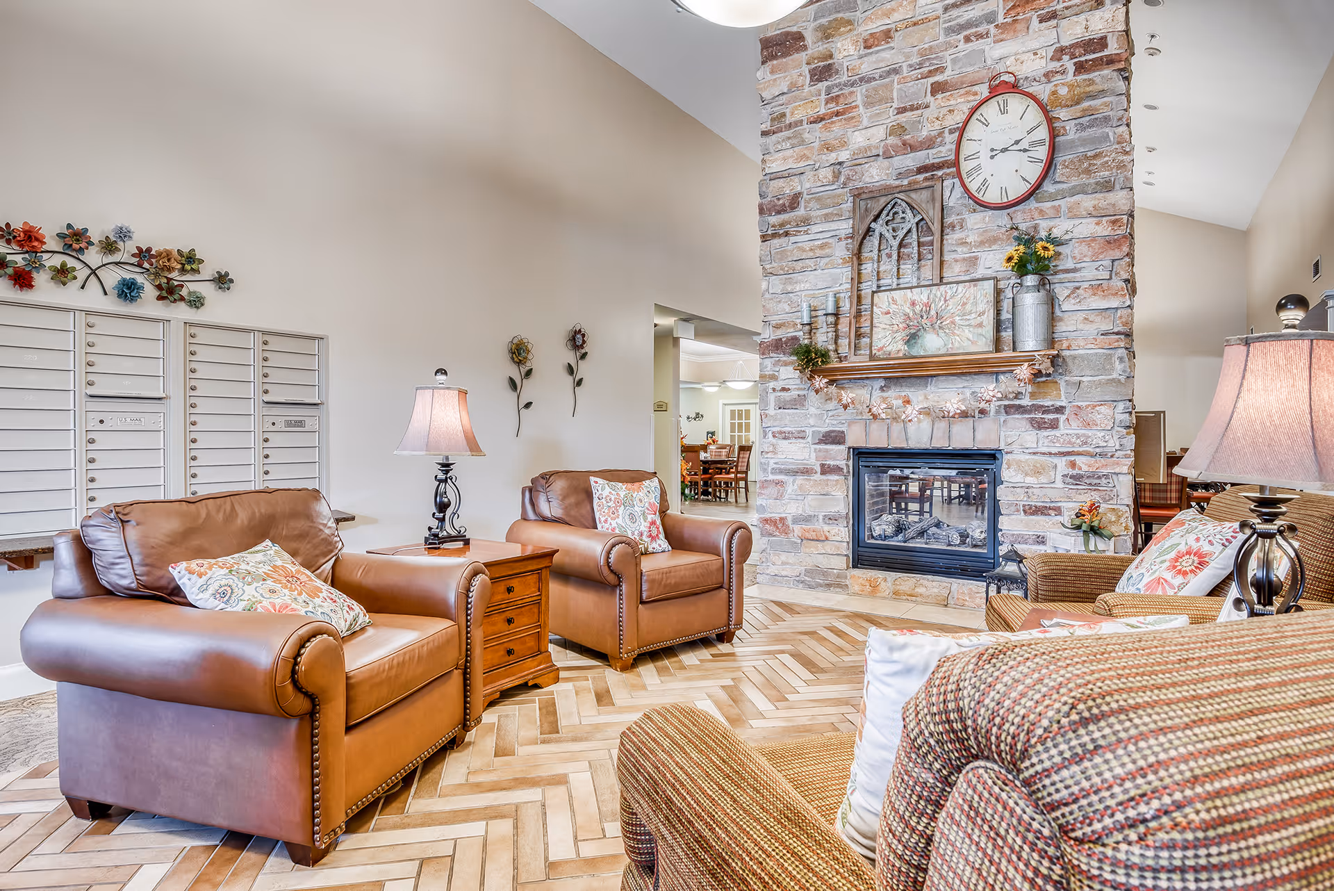 Common area lounge with leather armchairs, patterned cushions, a stone fireplace with a clock above, and mailboxes along the wall.
