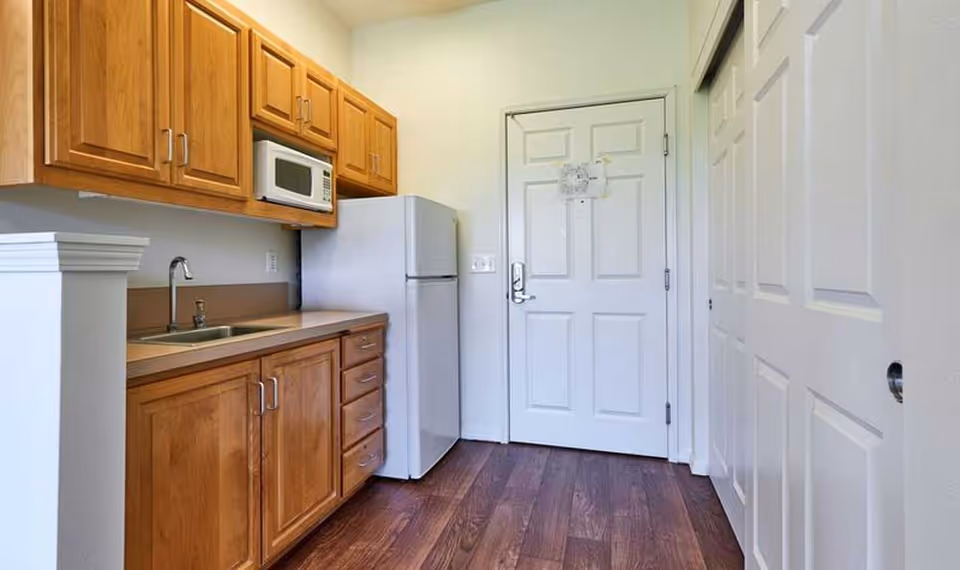 Small kitchenette with wooden cabinets, a sink, microwave, refrigerator, and a white entry door.