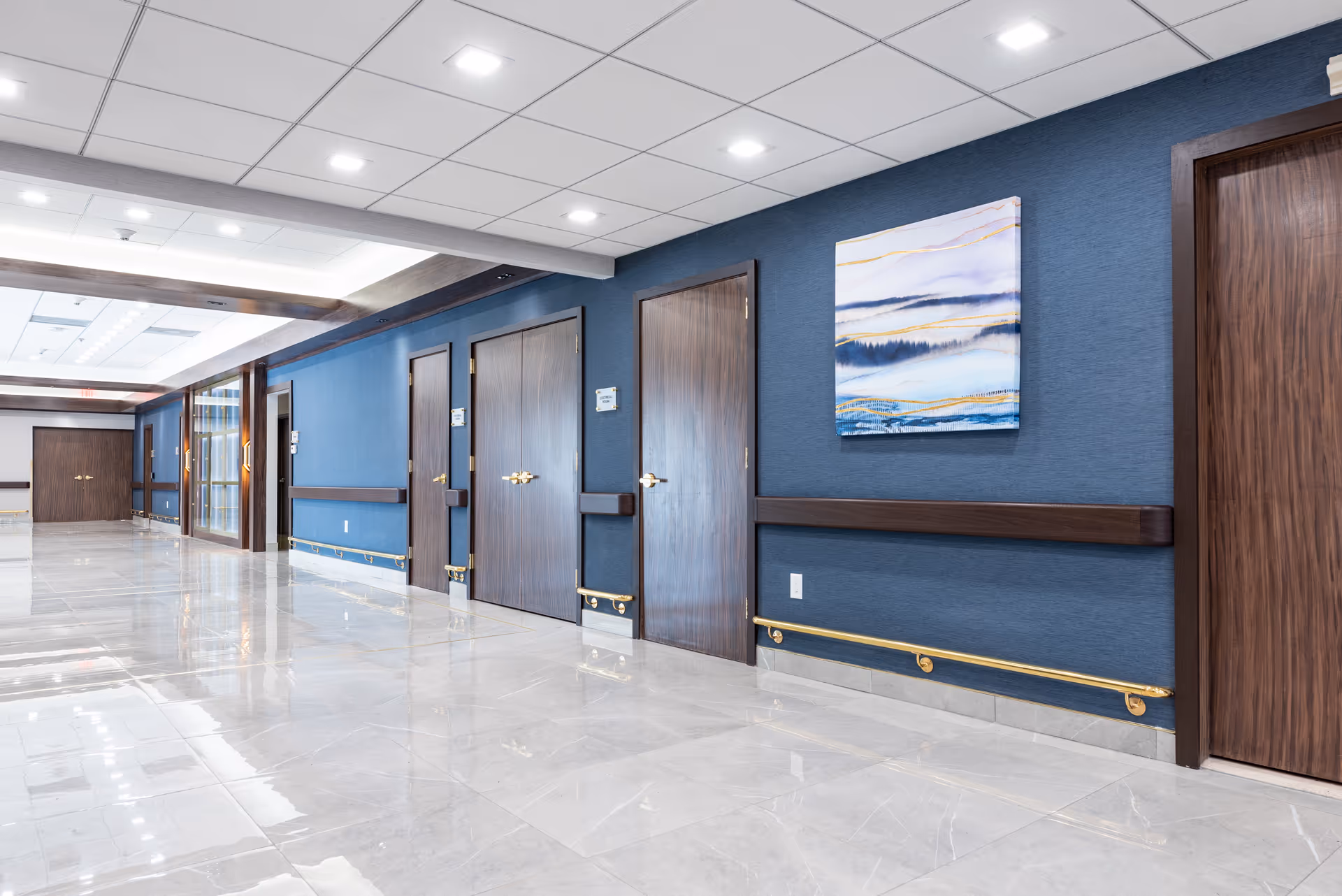 A clean, modern hallway in a senior living facility with polished tile floors, blue walls, several closed wooden doors with brass handles, handrails along the walls, and a framed abstract painting featuring blue and gold tones.