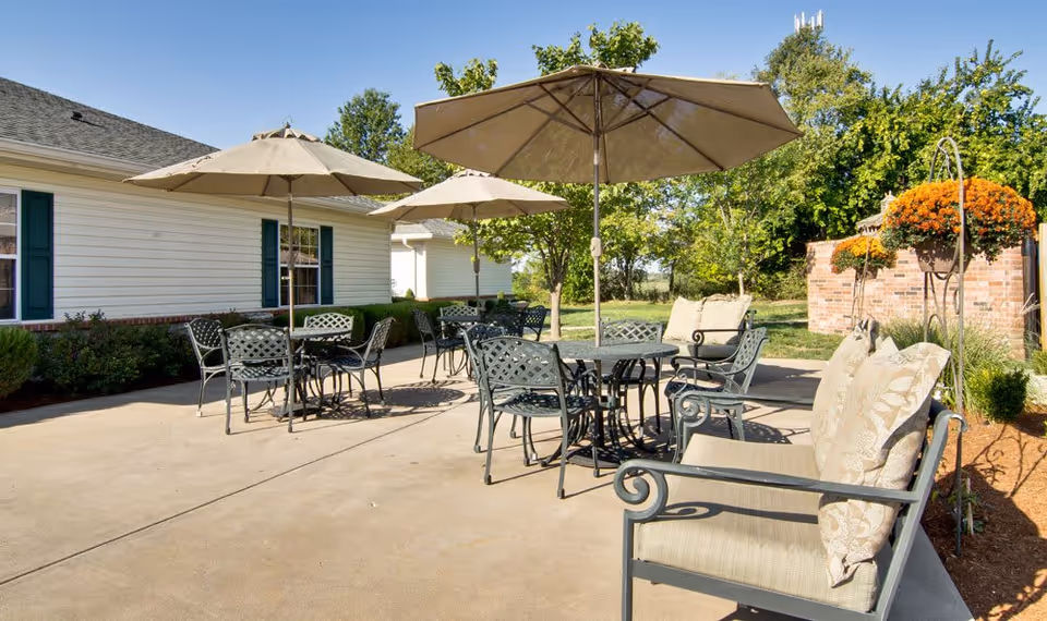 Outdoor patio area at Lakewood Senior Living with metal tables and chairs under large beige umbrellas, cushioned benches, potted orange flowers, and greenery surrounding the space on a sunny day.