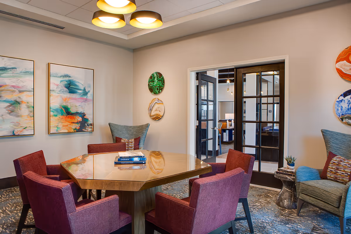 A bright communal dining room with a wooden hexagonal table surrounded by purple upholstered chairs, colorful wall art, and glass double doors.