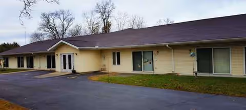 Single-story beige Rosebrooke Assisted Living building with front entrance, parking lot, and lawn.