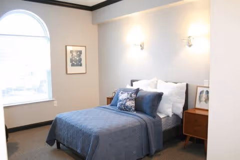 A neatly made bed with blue bedding and multiple pillows in a well-lit bedroom. There is a large arched window on the left side letting in natural light, a framed picture on the wall, and two wooden nightstands with lamps on either side of the bed.