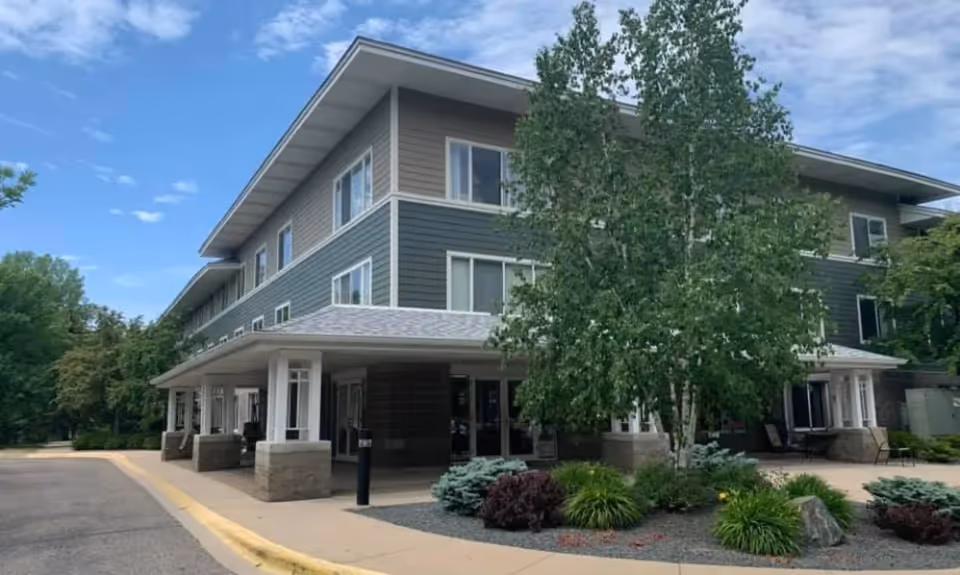 Exterior view of a three-story senior living facility building with a covered entrance, surrounded by trees and landscaped greenery under a partly cloudy sky.