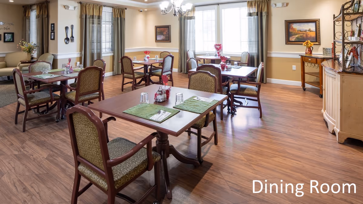 A well-lit dining room with several wooden tables and cushioned chairs arranged neatly. Each table is set with green placemats, utensils, glasses, and small decorative centerpieces. The room has wood flooring, beige walls with white trim, large windows with curtains, and framed artwork on the walls. There is a small sideboard with decorations on the right side of the room.
