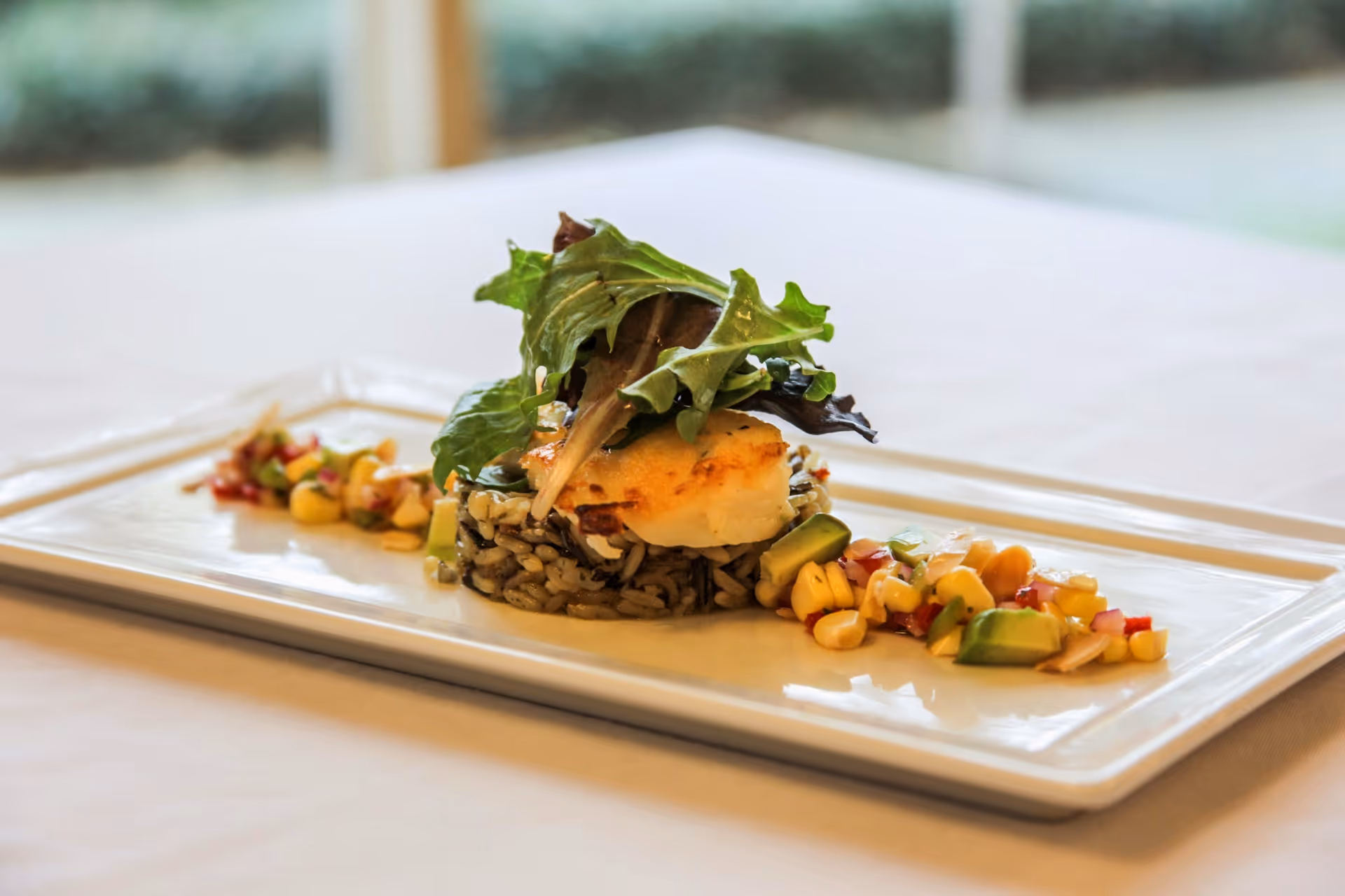 A gourmet dish served on a rectangular white plate featuring a portion of cooked rice topped with a piece of grilled fish and garnished with fresh leafy greens, accompanied by a side of corn and avocado salad.