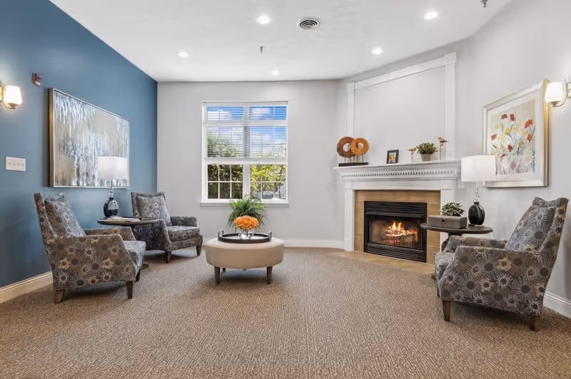 A cozy living room with a lit fireplace, four patterned armchairs arranged around a round ottoman with a flower arrangement, two side tables with lamps, artwork on the walls, and a window letting in natural light.