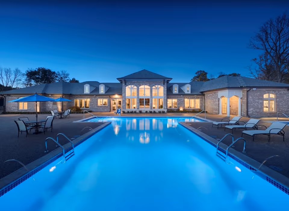 Evening view of a senior living facility with a large illuminated outdoor swimming pool in the foreground, surrounded by lounge chairs and tables with umbrellas. The building in the background has large windows with warm interior lighting and a pitched roof.