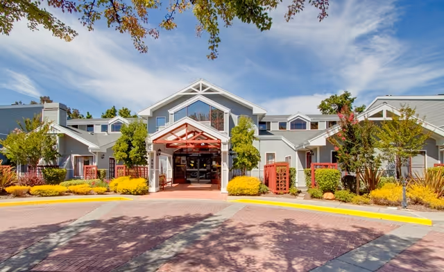 Front entrance of a two-story senior living building with a covered porte-cochère, landscaped shrubs, and a blue sky.