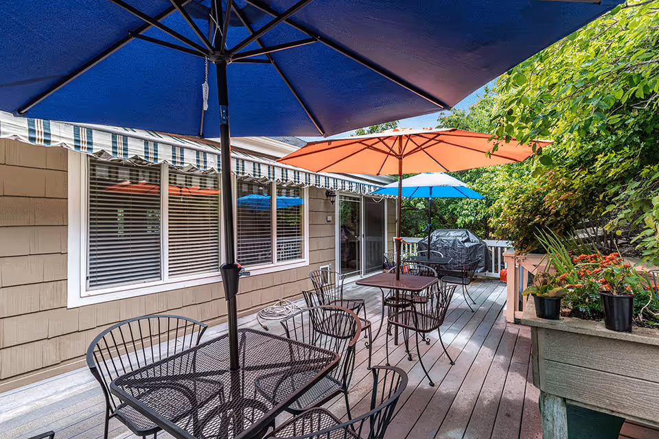 Outdoor patio area with multiple metal tables and chairs, each shaded by large colorful umbrellas in blue, orange, and light blue. The patio is adjacent to a building with windows and a striped awning. There are plants and greenery along the right side, and a covered grill is visible in the background.