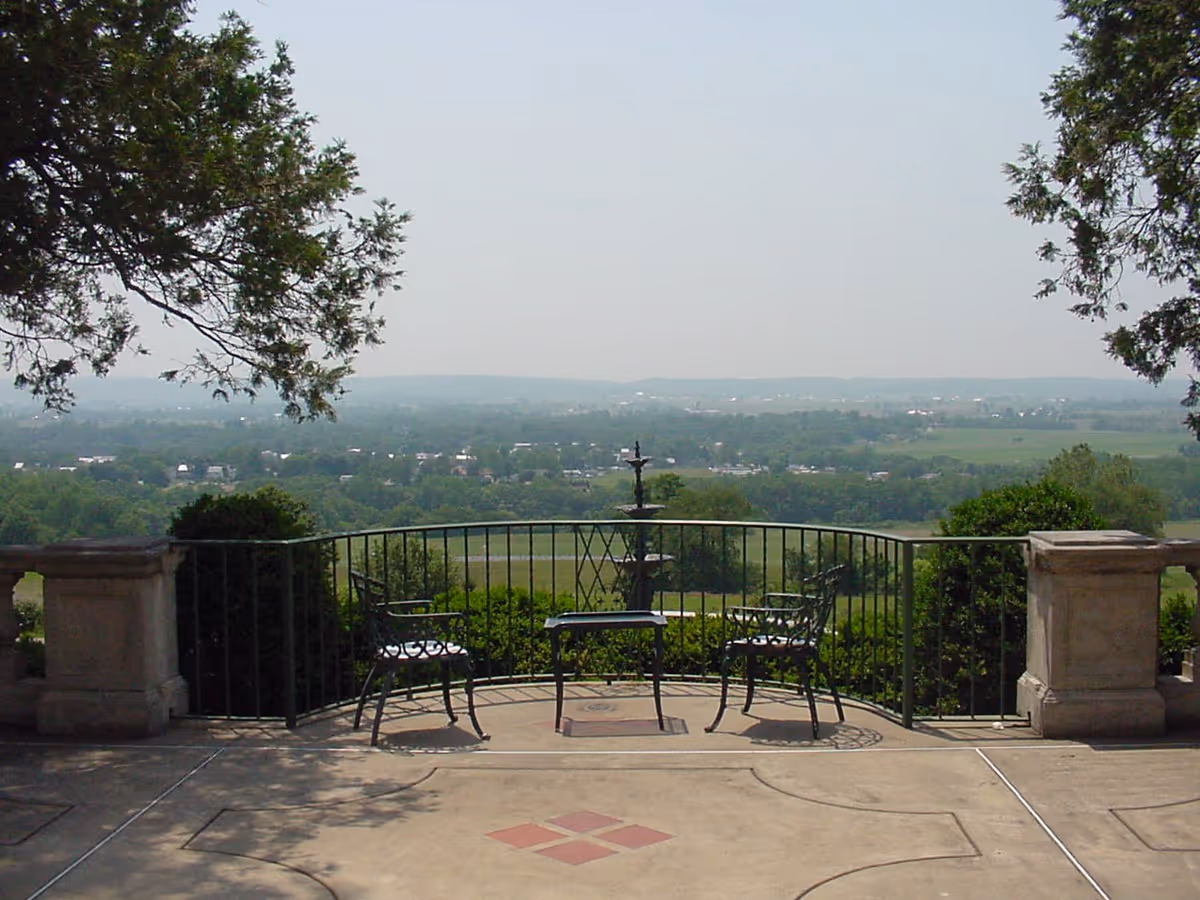 Outdoor patio area with two metal chairs and a small table facing a scenic view of a green landscape and distant hills, framed by trees and a decorative railing.