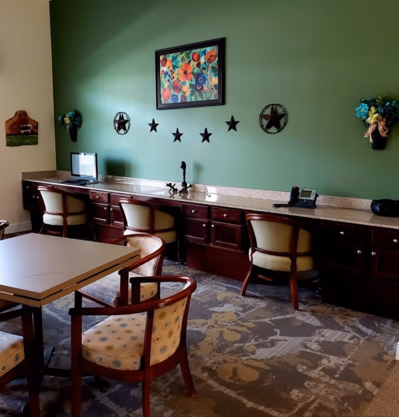 A room with a green accent wall decorated with a colorful floral painting and star-shaped wall hangings. Below the wall is a long wooden counter with three cushioned chairs. On the counter are a computer monitor, a telephone, and decorative items. In the foreground, there is a square table surrounded by four cushioned chairs with wooden frames. The floor is covered with a patterned carpet.